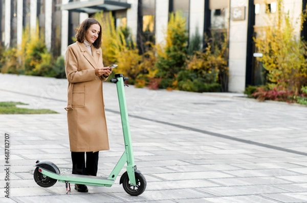 Fototapeta Happy woman in autumn coat using smartphone for renting modern electric scooter with an application, reads qr code with a mobile phone camera. Alternative city transport