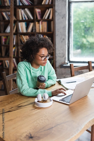 Fototapeta african american student in eyeglasses using laptop while holding paper cup near headphones on desk