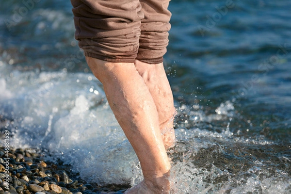 Fototapeta The legs of an elderly woman with altered varicose veins on the background of the sea in autumn. Lifestyle, tempering fall.
