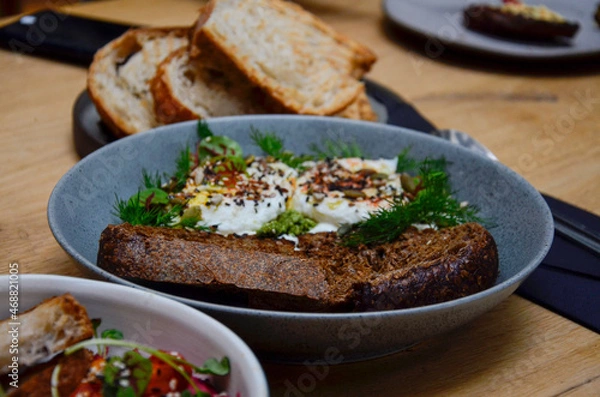Fototapeta On the table in a plate is a slice of rye bread, poached eggs sprinkled with cereal seeds and lined with dill sprigs. Behind her is a plate of toasted white bread.