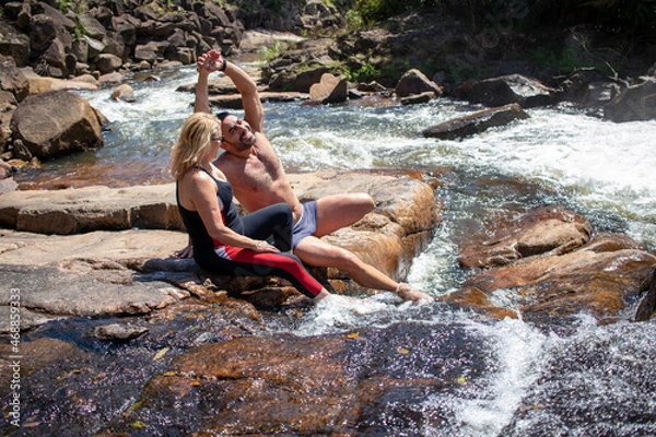 Obraz happy couple talking and relaxing, sitting on rock with river water in sunny day.