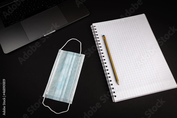 Fototapeta Blank notebook with pencil on black background with laptop computer. Medical mask laying next to notebook. Flat lay image for business concept.