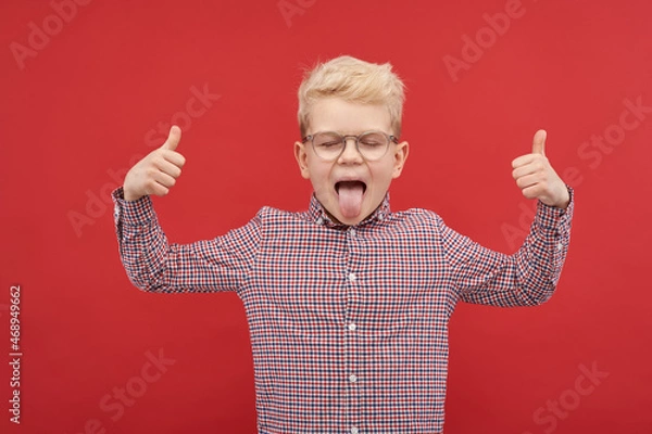 Fototapeta Emotions, good mood, vision test, health project. Happy child shows tongue and thumb up gesture. Portrait of a boy in a plaid shirt and glasses. Back to school.