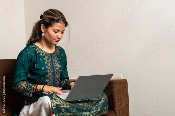 Fototapeta Happy indian girl relaxing on comfortable couch and working on laptop at home. Surfing internet and work from home concept.