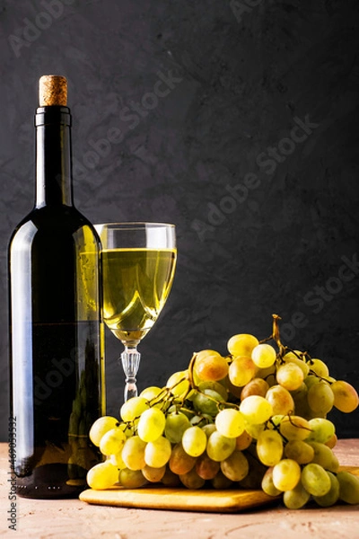 Obraz a glass glass and a bottle of white wine and a brush of white grapes on a cutting board on a black background
