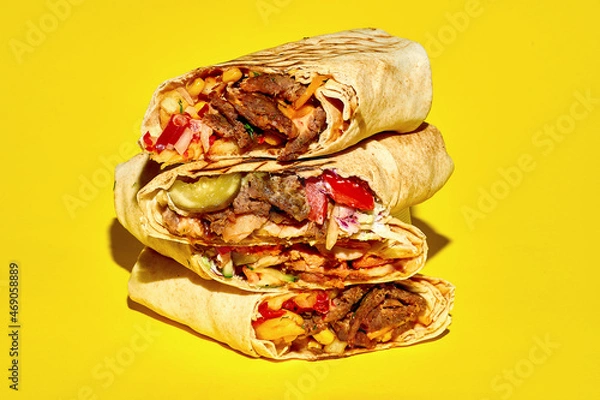 Fototapeta A stack of different shawarma with meat, vegetables and falafel on a yellow background. Close-up, selective focus