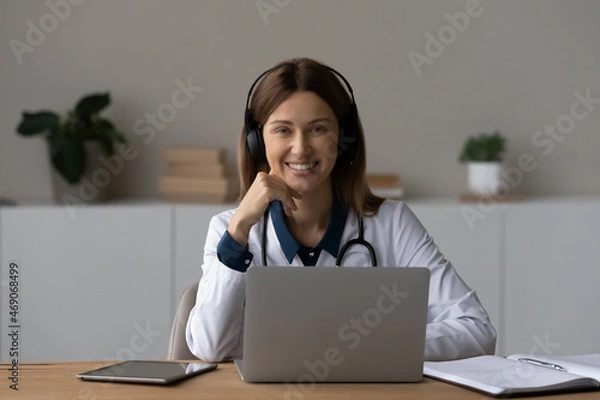 Fototapeta Head shot portrait smiling female doctor therapist in wireless headphones sitting at work desk with laptop, professional practitioner gp looking at camera, consulting patient online by video call