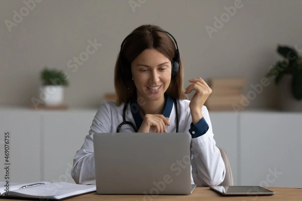 Fototapeta Smiling woman doctor in headphones consulting patient online, looking at laptop screen, young female practitioner therapist gp making video call or watching webinar, sitting at work desk in office