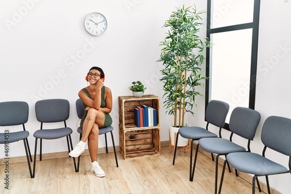 Fototapeta Young hispanic woman listening to music sitting on chair at waiting room