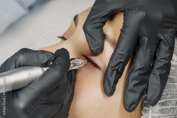 Fototapeta Permanent make up eyeliner procedure, applying on young girl. Close-up. Young beautiful woman making permanent makeup in cosmetology salon. Young girl applying permanent eyeliner in beauty studio