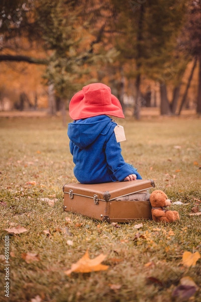 Fototapeta A child in a blue coat and a red hat is sitting on a suitcase. A children's photo shoot in the image of Paddington bear