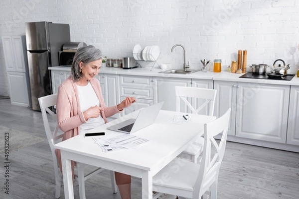 Fototapeta Cheerful mature woman pointing with fingers during video call on laptop near paper with bills in kitchen.