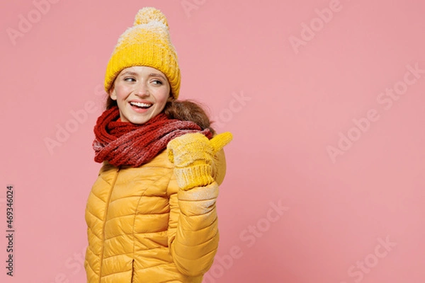Fototapeta Promoter smiling fun young woman 20s years old wears yellow jacket hat mittens pointing back behind on workspace area copy space mock up isolated on plain pastel light pink background studio portrait.