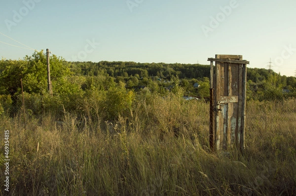 Fototapeta An old wooden door standing in the middle of the road among trees and bushes on a clear cloudless summer evening