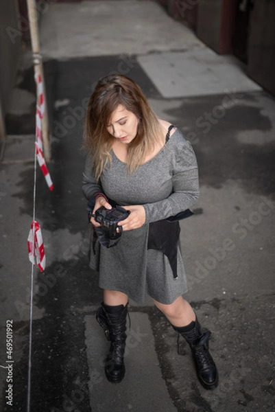 Fototapeta A curvy photographer girl in a gray short dress, leather platform boots and with a camera in her hands looks through the material taken at the photo shoot on the camera screen