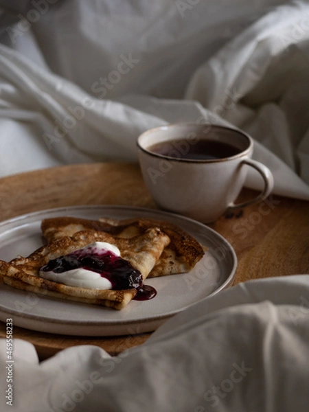 Fototapeta Breakfast in bed, a cup of black tea and thin pancakes with currant jam and sour cream on a beige ceramic plate on a wooden tray on a soft white bed in the morning light