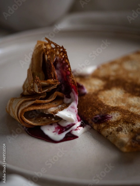 Fototapeta Breakfast in bed, a cup of black tea and thin pancakes with currant jam and sour cream on a beige ceramic plate on a soft white bed in the morning light