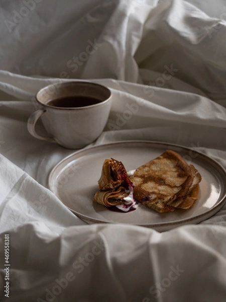 Fototapeta Breakfast in bed, a cup of black tea and thin pancakes with currant jam and sour cream on a beige ceramic plate on a soft white bed in the morning light