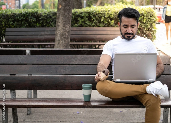 Fototapeta Latin man using his laptop on a plaza bench with a coffee mug
