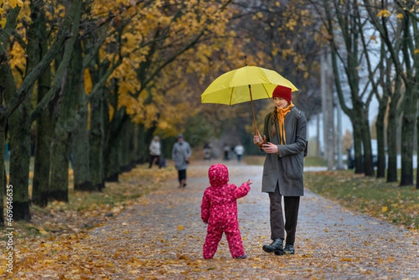 Fototapeta Mom walks with small child in an autumn park under a yellow umbrella