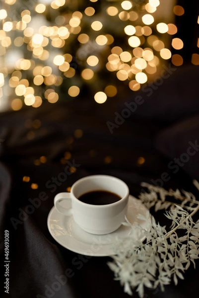 Fototapeta White cup of coffee on a dark bed with pillows in a cozy room with a christmas tree and blurred lights