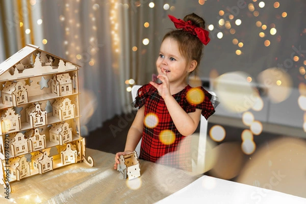 Fototapeta A smiling  pretty girl with sweets in hand, finding gift in toys wooden house of Christmas Advent calendar, month numbers of December. Concept children fun, waiting for a miracle