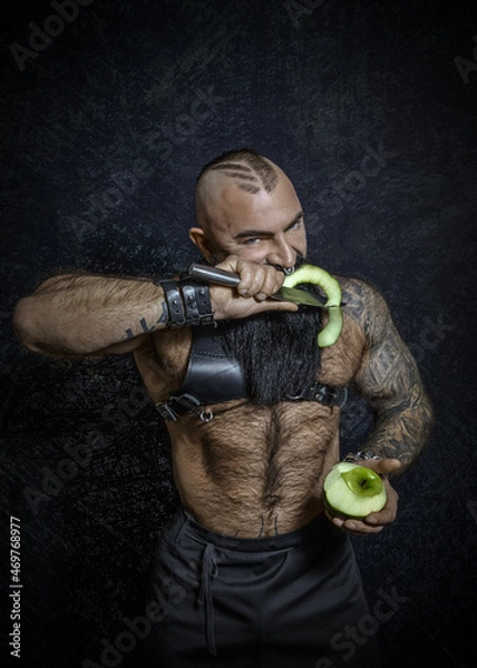 Fototapeta A brutal sexy man with a black beard with a naked hairy torso cuts a green apple with a large knife and eats a piece of apple