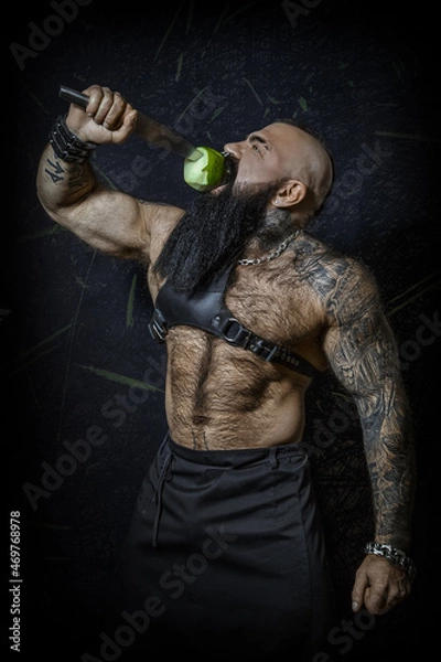 Fototapeta Brutal sexy man with a black beard with a naked hairy torso bites a green apple on a large knife