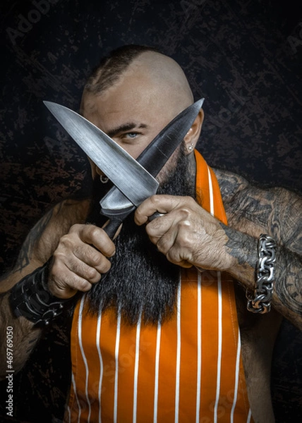 Fototapeta A brutal sexy man with a black beard in an apron has crossed big knives and looks menacing