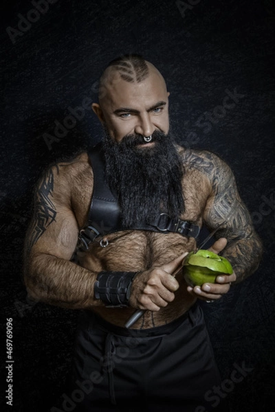 Fototapeta A brutal sexy man with a black beard with a naked hairy torso cuts a green apple with a large knife