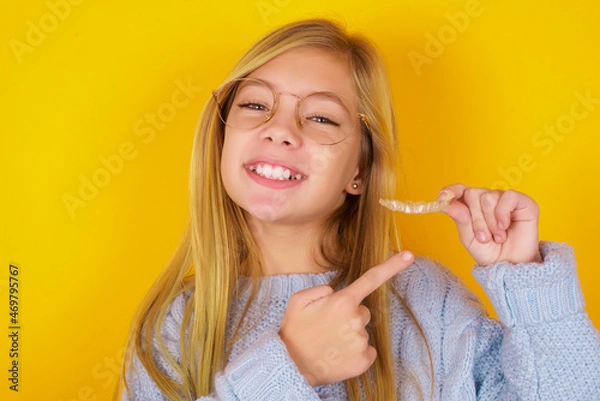 Fototapeta caucasian kid girl wearing blue knitted sweater over yellow background  holding an invisible aligner and pointing at it. Dental healthcare and confidence concept.