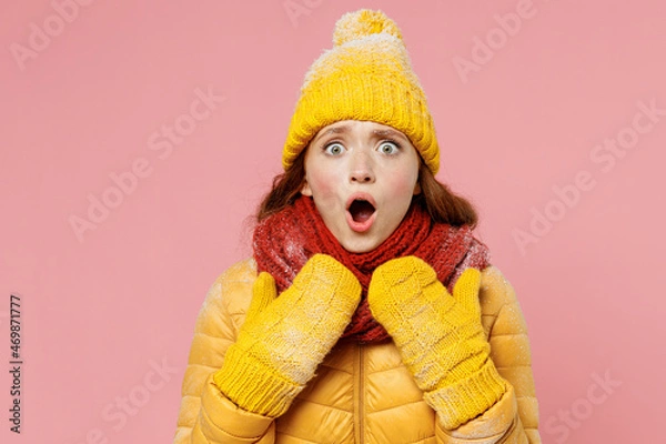 Fototapeta Frightened afraid scared amazed young woman 20s wears yellow jacket hat mittens look camera keeping mouth wide open point hands herself isolated on plain pastel light pink background studio portrait.