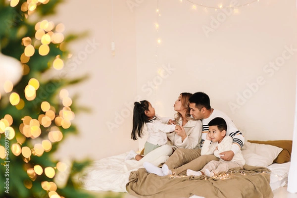 Fototapeta A mixed race Asian family with two children in cozy sweaters is relaxing together, resting and having fun on a bed in a decorated bedroom with Christmas tree at home in the New Year. Selective focus