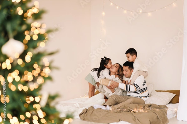 Fototapeta A mixed race Asian family with two children in cozy sweaters is relaxing together, resting and having fun on a bed in a decorated bedroom with Christmas tree at home in the New Year. Selective focus