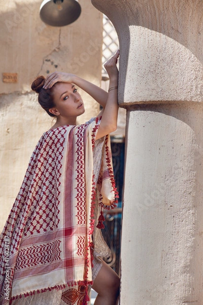 Obraz red-haired beautiful girl in Arabic clothes stands against the background of the old arabic city, bazaar. dressed in a beige abaya with embroidery and a scarf on her head