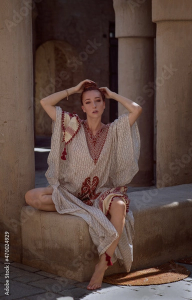 Obraz red-haired beautiful girl in Arabic clothes sit against the background of the old arabic city, bazaar. dressed in a beige abaya with embroidery and a scarf on her head