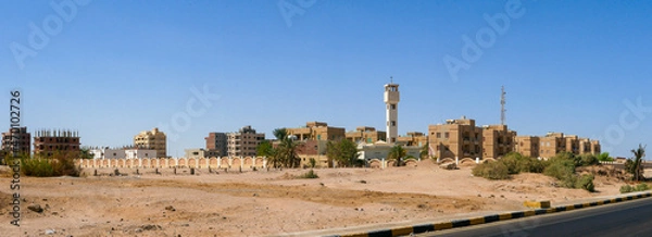 Fototapeta Panoramic view from the road to the low houses of local residents surrounded by desert and palm trees against the background of a blue sky. Copy space. Safaga, Egypt.