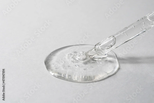 Fototapeta Facial serum pouring from glass pipette on grey textured background. Transparent liquid gel drop with bubbles closeup. Skin care routine. Trendy cosmetic product macro. Front view.