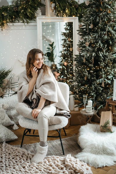 Obraz Overjoyed young happy european woman in warm knitted sweater sitting under decorated Christmas tree, holding mobile call conversation with relatives or friends, congratulating telling best wishes.