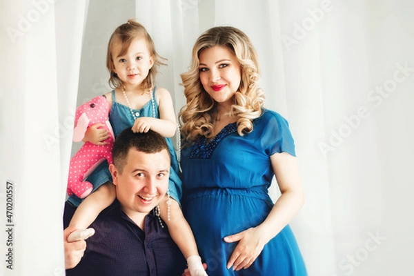 Obraz portrait of a family. a little daughter sits on her father's neck, mother stands nearby. The woman is pregnant. All dressed in blue