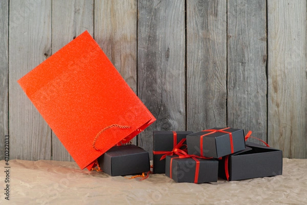 Fototapeta Gifts in black boxes tied with a red ribbon and a red shopping bag stand on craft paper against a wooden background