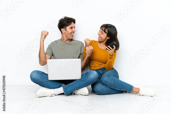 Fototapeta Young couple with a laptop and mobile sitting on the floor celebrating a victory in winner position