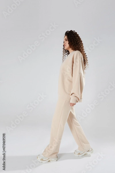 Obraz Young woman with afro hairstyle in modish sweatshirt and pants walking across a white background