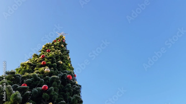 Fototapeta An elegant lonely Christmas tree decorated with garlands and a star against the blue sky. Outdoors, green decorated Christmas tree. As a background and texture for a holiday card with a place for text