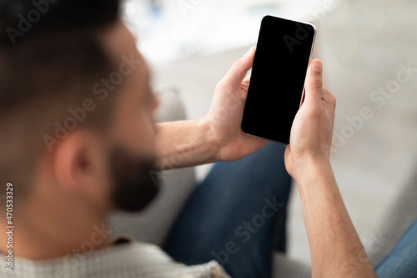 Fototapeta Unrecognizable young Arab man using smartphone on sofa at home, mockup for app or website design