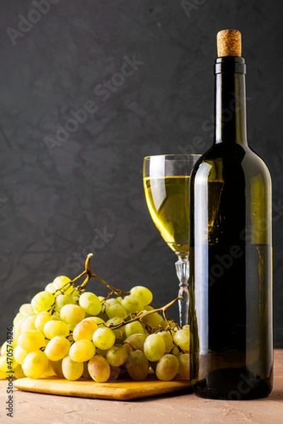 Obraz a glass glass and a bottle of white wine and a brush of white grapes on a cutting board on a black background
