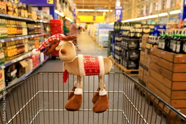 Fototapeta Funny Christmas deer on supermarket trolley. Shopping in the shop before the New Year, sale. A basket in wholesale store, background of blurred shelves with groceries and goods.