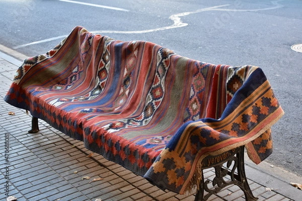Fototapeta The street bench is covered with a beautiful colored carpet. A bench with a cast-iron base on the street of Yerevan.