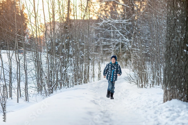 Fototapeta A cheerful and handsome boy in blue clothes runs along a snow-covered path in the park. Winter activities.