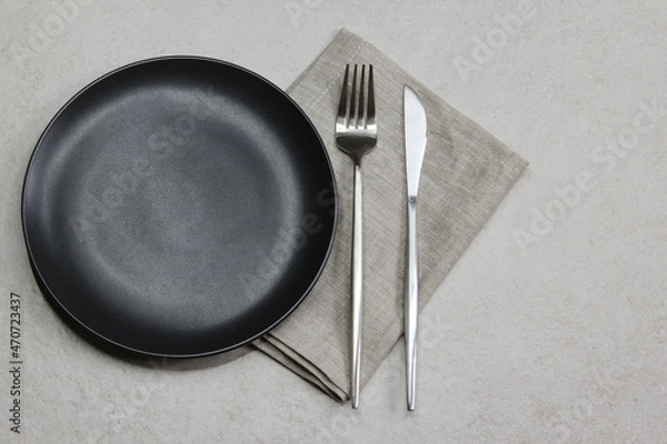 Fototapeta Black empty plate with knife and fork with gray textile napkin on grey stone background. Table setting. Top view. 
